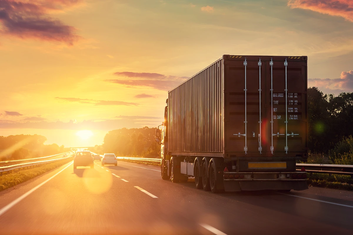 Container truck driving on a highway at sunset with cars on the road.