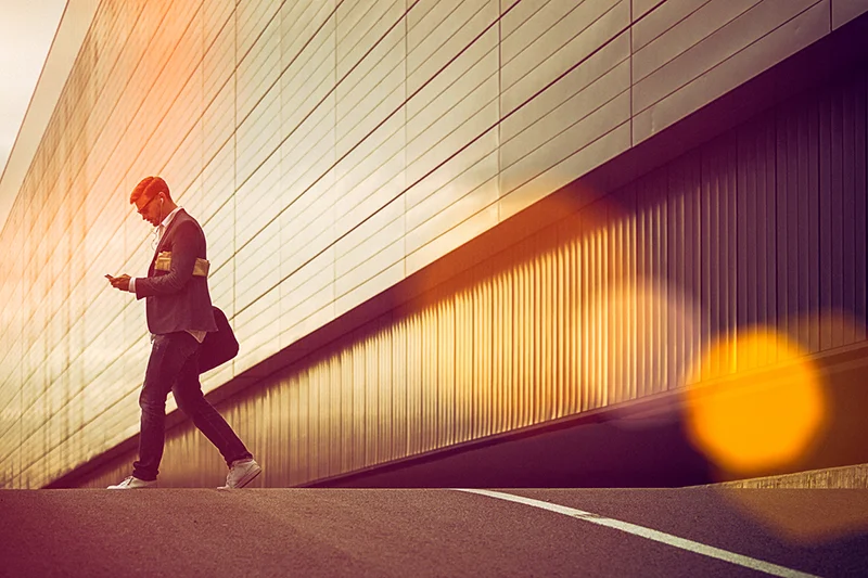 Man walking beside a modern building while looking at his phone in warm sunlight