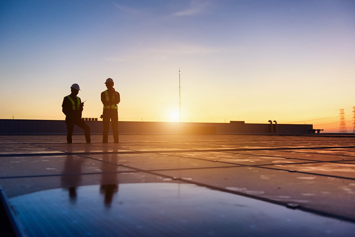 Two workers in hard hats standing on a rooftop at sunset.